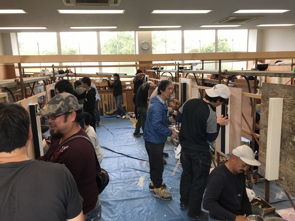 ブルーシートの敷かれた上の左官練習台にむかって作業を進める複数の男性と見守る久住さん