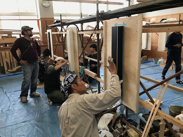 ブルーシートの敷かれた上の左官練習台にむかって作業を進める男性とそれを見守る男性