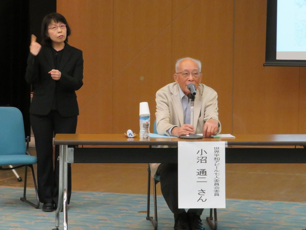 小沼先生が椅子に座り放されていて同時に手話の方が後方で通訳されている様子の写真