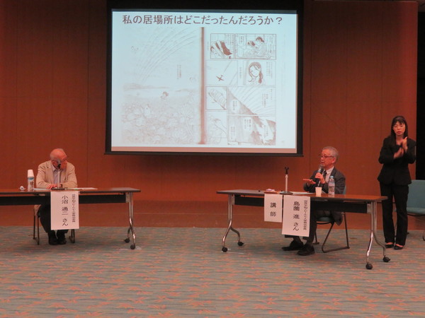 スクリーンの前で小沼先生と島薗先生が椅子に座りながら対談されていて同時に手話通訳されている様子の写真