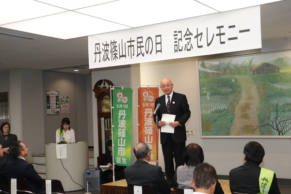 市民ホールにて丹波篠山市民の日記念セレモニーと書かれたプレートの下で椅子に座って聞く観客にむけ話す市長の様子