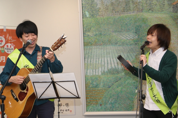 舞台上でそれぞれギターとタンバリンをもってマイク前で歌うふるさと大使のちめいどのお二人の写真