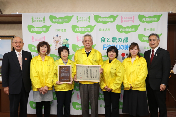 丹波篠山市と書かれた壁の前で、酒井市長、前川教育長、黄色いジャンパーを着たきたっこ守り隊の方々が賞状をもって笑顔で立っている写真