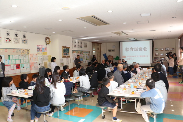 机に並べられた給食を前にそれぞれ椅子に座り、スクリーンを見ながら説明を受けている様子