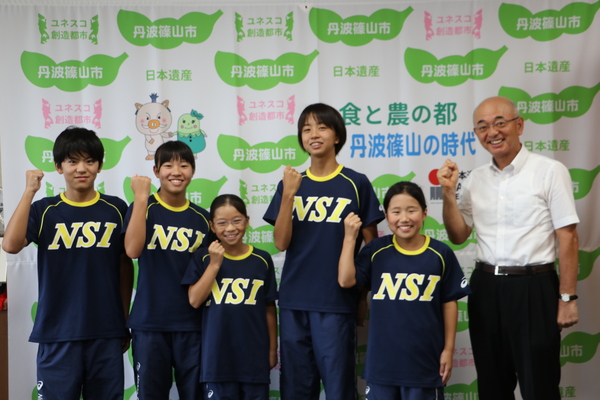 丹波篠山市と書かれた壁の前で、市長とNSIと書かれたユニフォームを着た子どもたちがガッツポーズをして笑顔で立って並んでいる写真