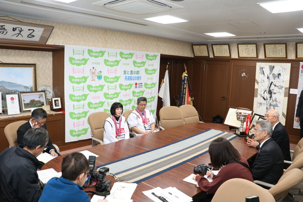 テーブルを囲み、椅子にかけて新聞記者やインタビューアーに説明するメダルをかけた栄養教諭と調理員さん