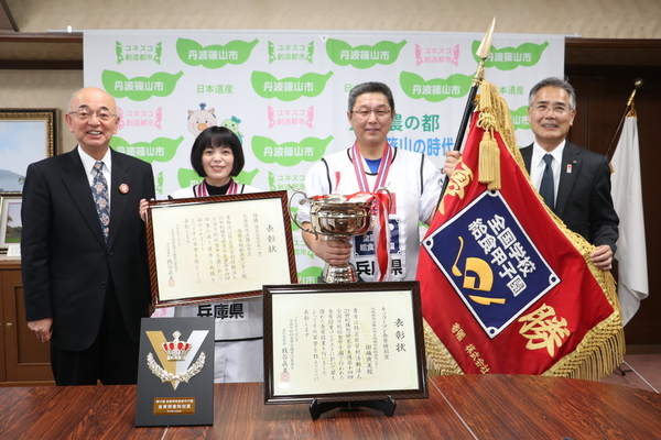 丹波篠山市と書かれた壁の前で、表彰状や大きなトロフィー、優勝旗を持った栄養教諭田畑さんと調理員出野さん、酒井市長と前川教育長の写真