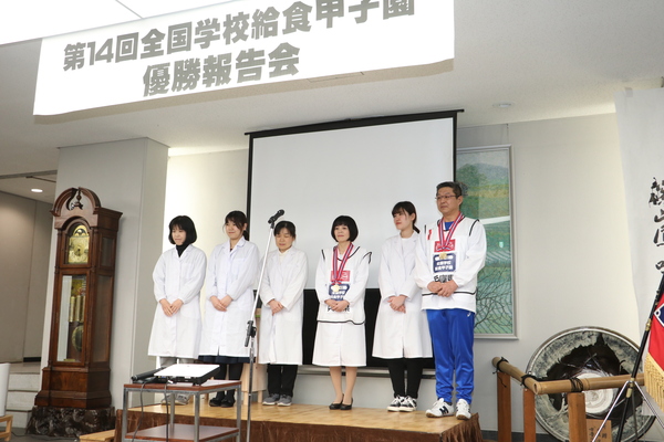 第十四回全国学校給食甲子園優勝報告会のパネル下で表彰される給食センター員の皆さん