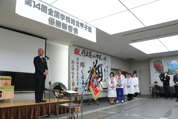 市民ホールにて舞台マイク前で話す酒井市長と、舞台横でそれを聞く表彰者の給食センター員の皆さんの写真