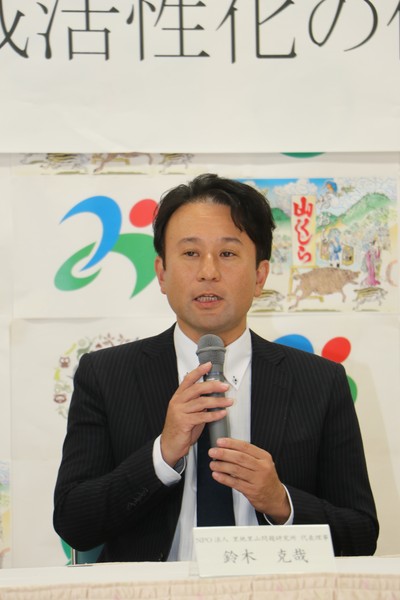 マイクを持ちスーツ姿で話をする、さともんの鈴木さん