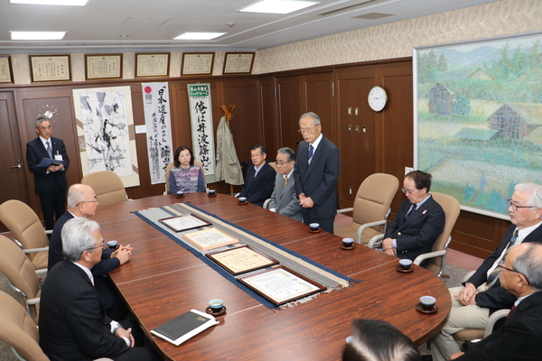 応接室にて大きなテーブルを囲み椅子に座り話を聞く市長、表彰者の方々と、立ってお話される表彰者の男性