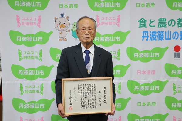 ともしび賞の賞状を両手で持ち、丹波篠山市と書かれた壁の前で立つ小林さん