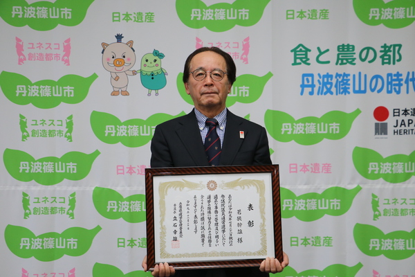 表彰状を両手で持ち、丹波篠山市と書かれた壁の前で立つ若狭さん