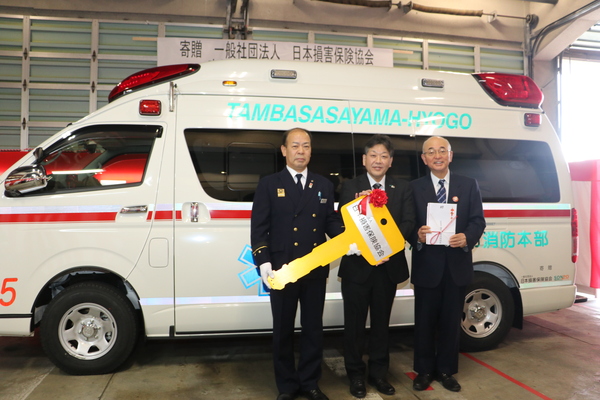 贈られた救急車の前で大きなリボンのカギを持った男性二名と市長