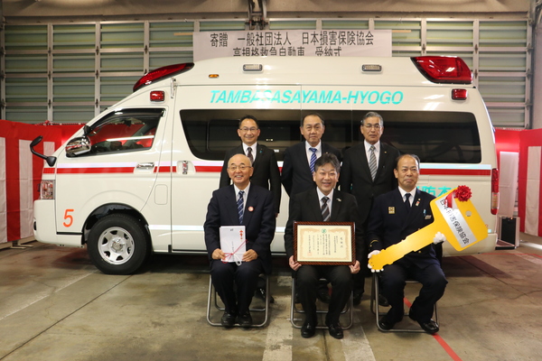 贈られた救急車の前で大きなリボンのカギと賞状を持った男性五名と市長