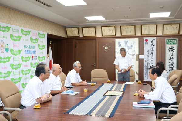 男性が向かい合って座り正面の男性が起立して文章を読んでいる写真