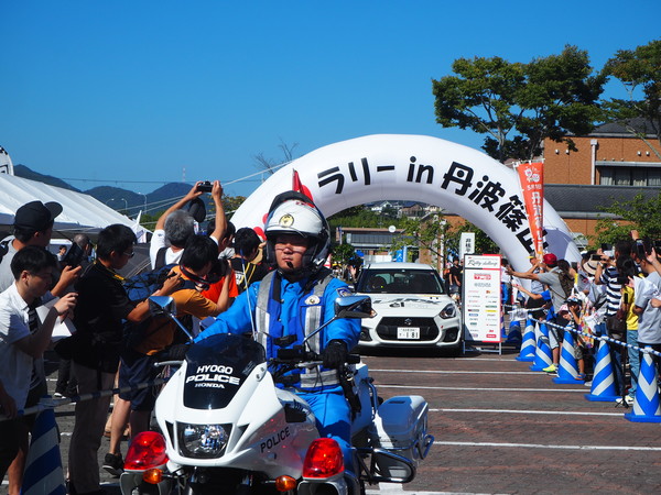 バイクの先導され、白いスポーツカーがゲートから出発する写真