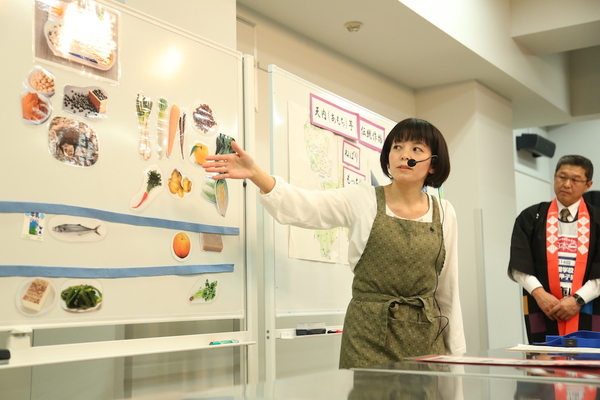 野菜や給食メニューの貼られたホワイトボードを前にマイクをつけて手で指し示しながら説明をする女性の写真