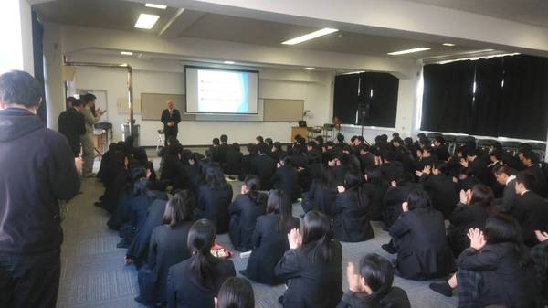 高校生が床に座り舞台のスクリーン前で話す市長にむけて拍手をおくっている様子