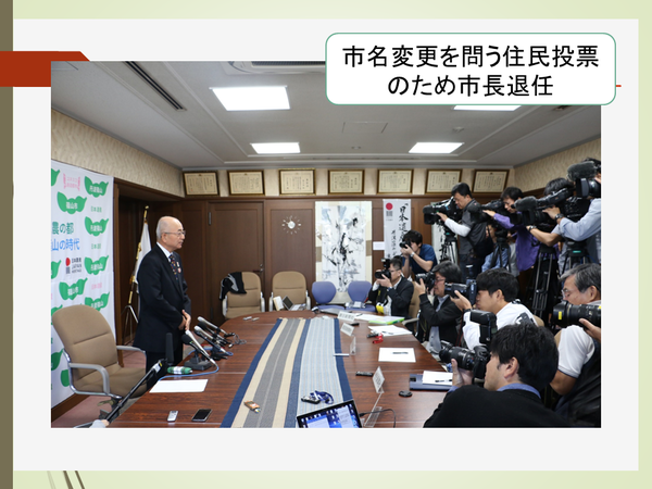 市名変更を問う住民投票のため市長退任という文字のうしろに、マイクの前で大勢のカメラマンや記者に向け、立って話す酒井市長