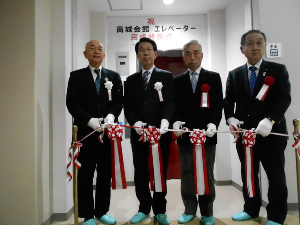 完成したエレベーター前でテープカットをするスーツ姿の四名の男性