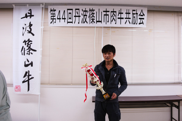 第四十四回丹波篠山市肉牛共励会のパネルの下でトロフィーをもって笑顔で立つ男性の写真