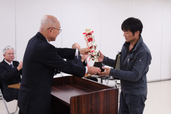 酒井市長から大きなトロフィーを受け取る男性と見守る椅子に座り拍手をおくる数人の男性