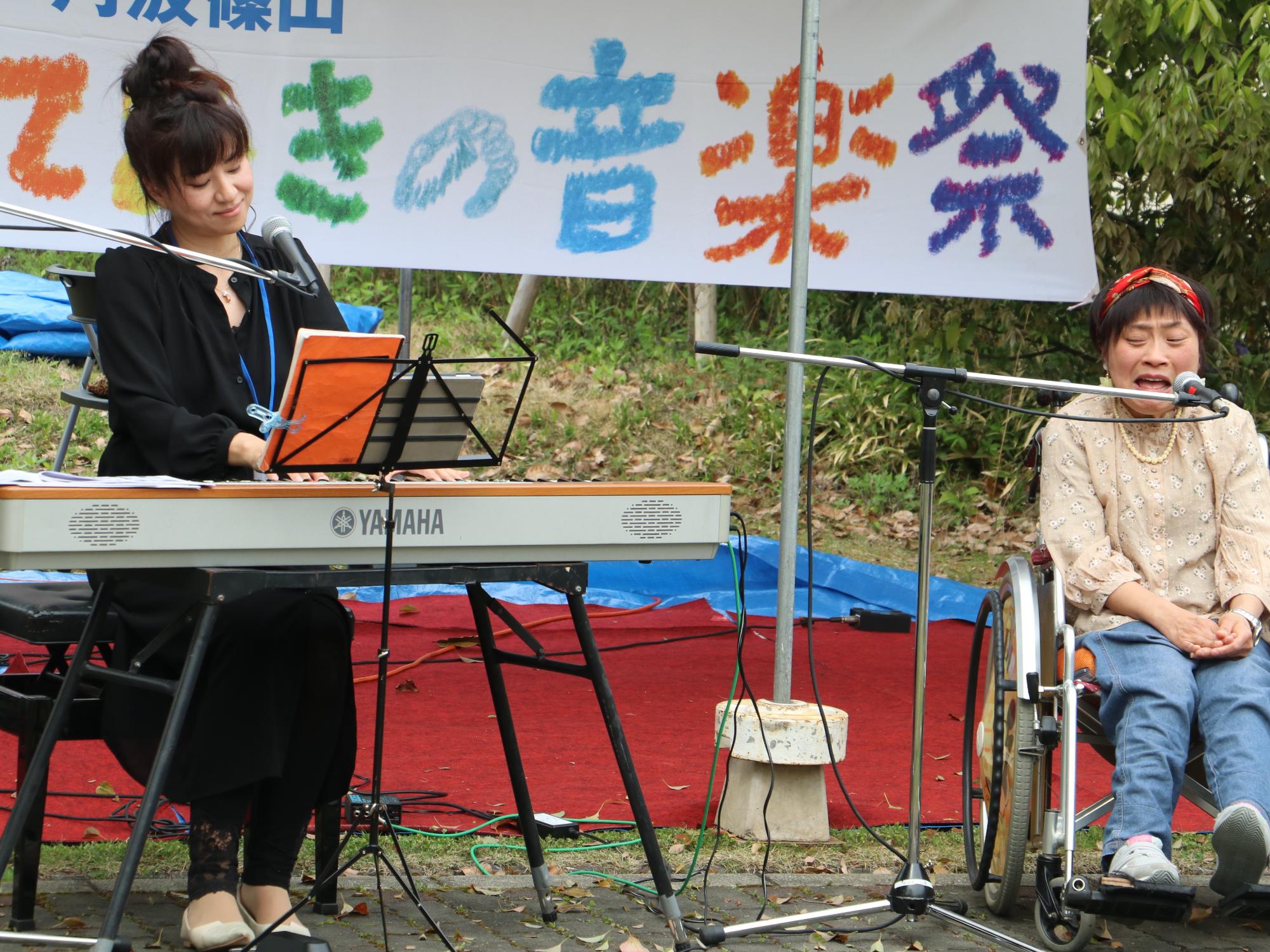 女性がキーボードを弾きながら歌い、その隣で女性が座って歌っている
