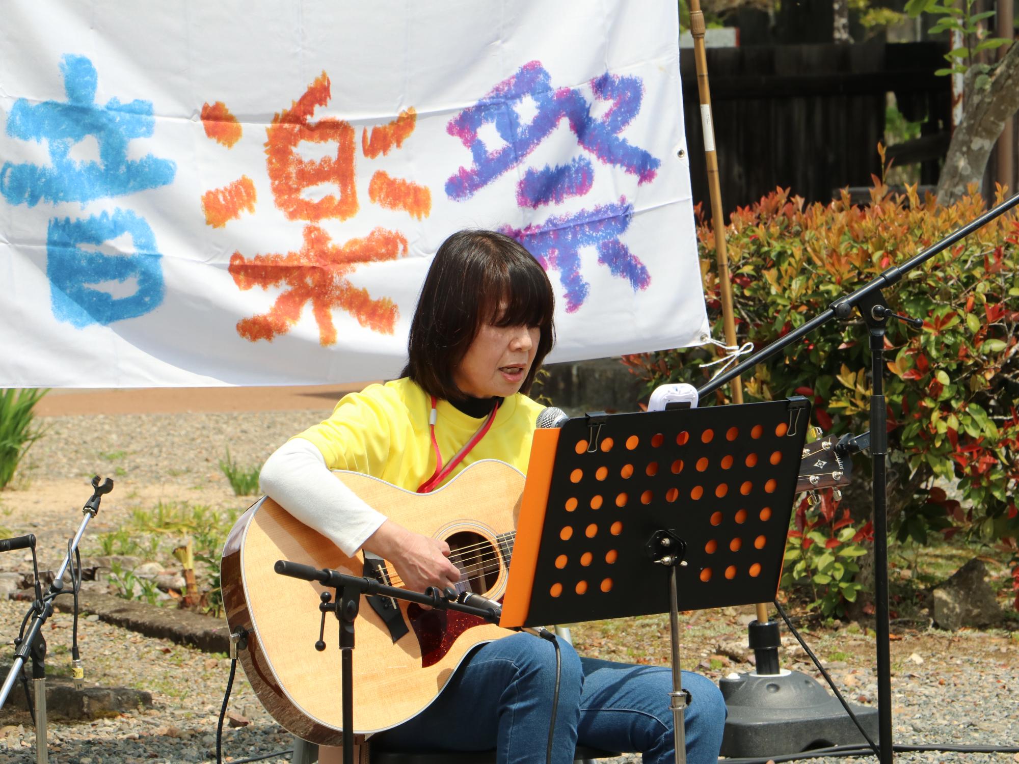 女性が座ってギターを弾きながら歌っている