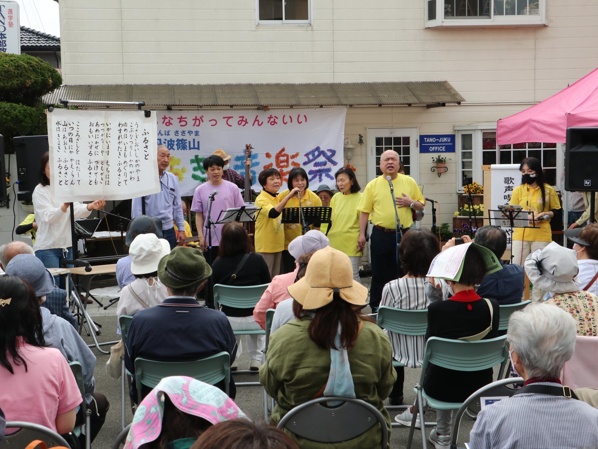 協会の前で歌声喫茶ささやまの皆さんが歌っている。パイプ椅子に座ってたくさんのお客さんが聴いている