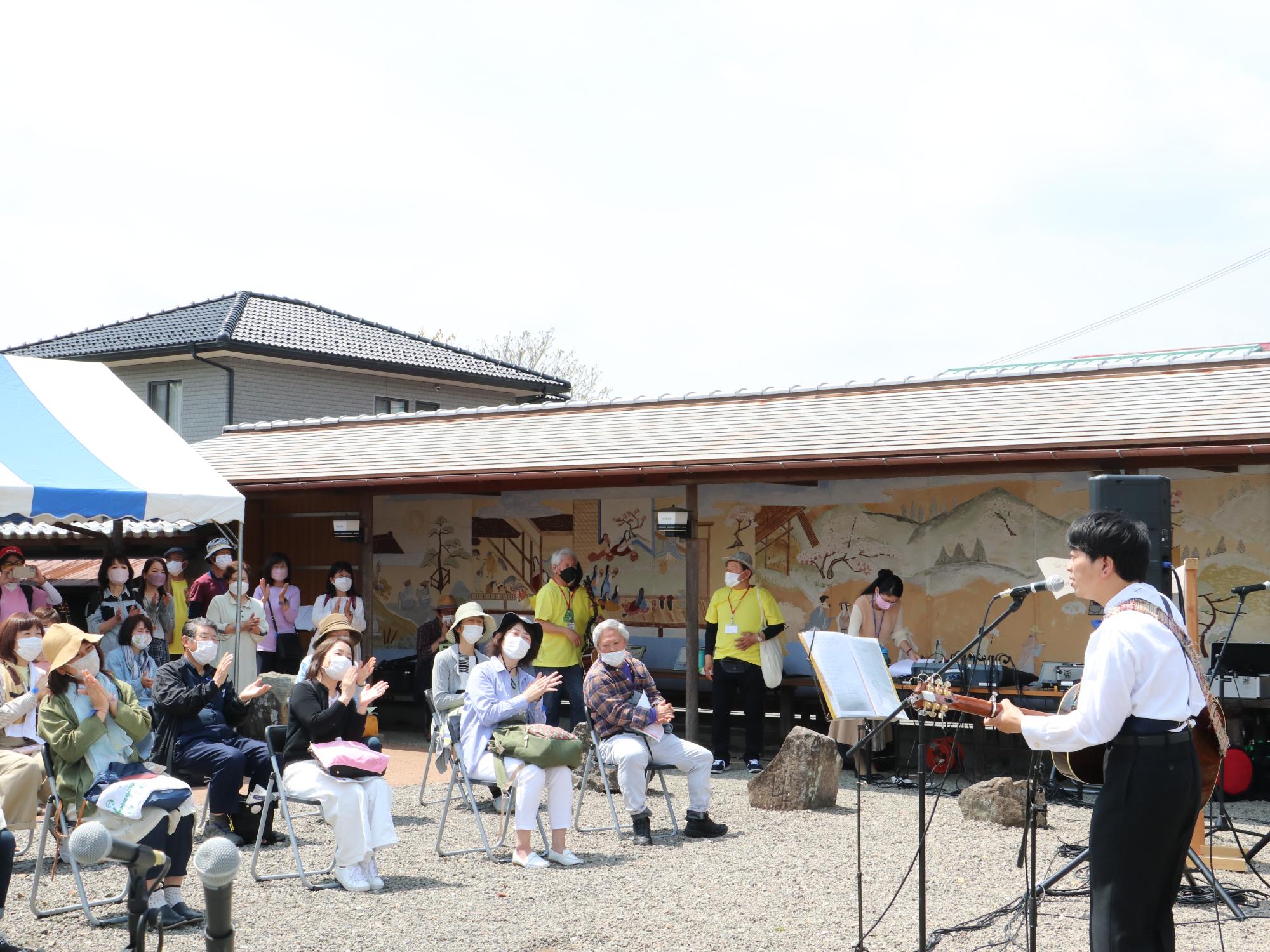 ちめいどの男性がギターを弾きながら歌っている。椅子に座ったお客さんが手拍子している