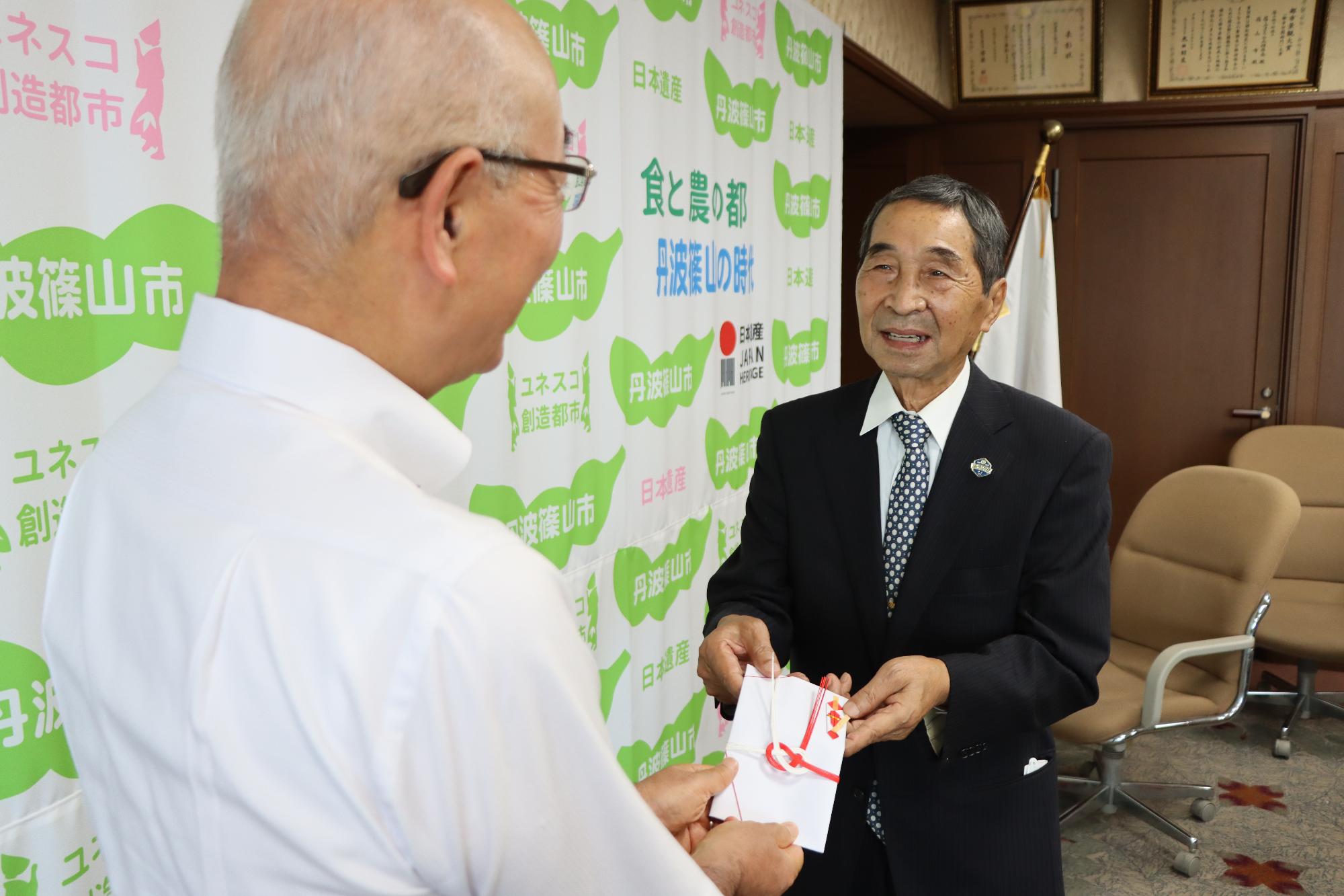 スーツを着た男性が、市長に目録を手渡ししている