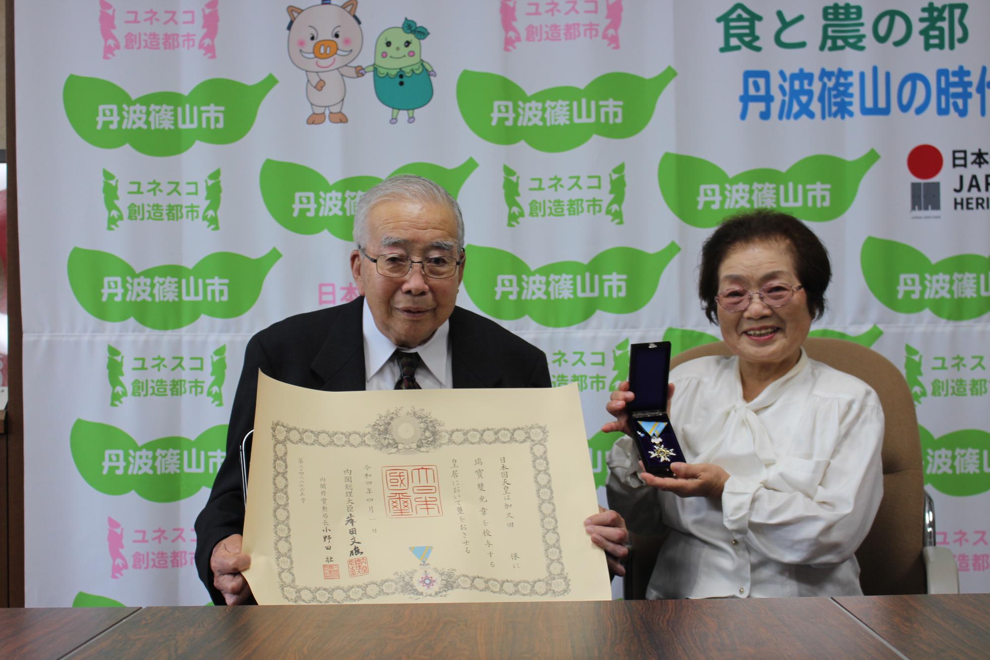 高齢者叙勲の賞状を受賞された男性が持っていて、その隣に奥様が勲章を持って座っている