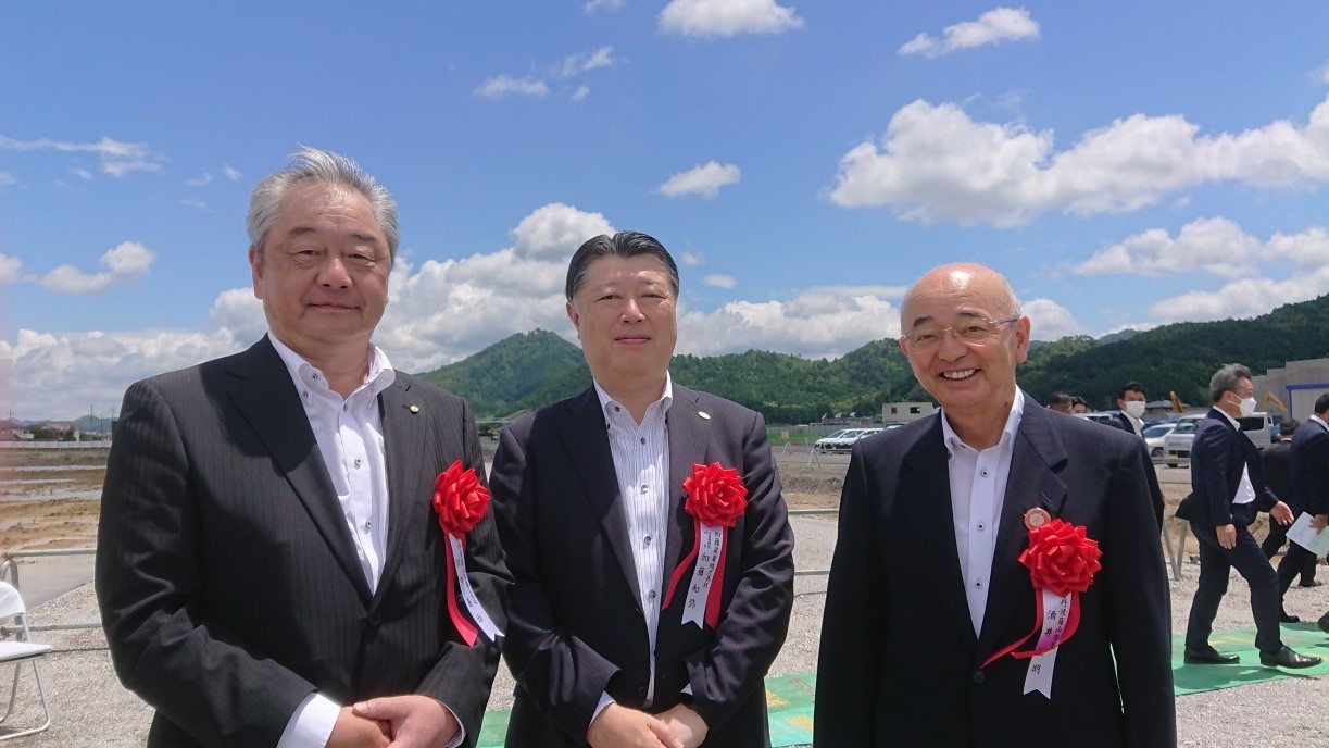 スーツを着て、胸元に赤いリボンをつけた男性3人が、高城山を背に記念撮影