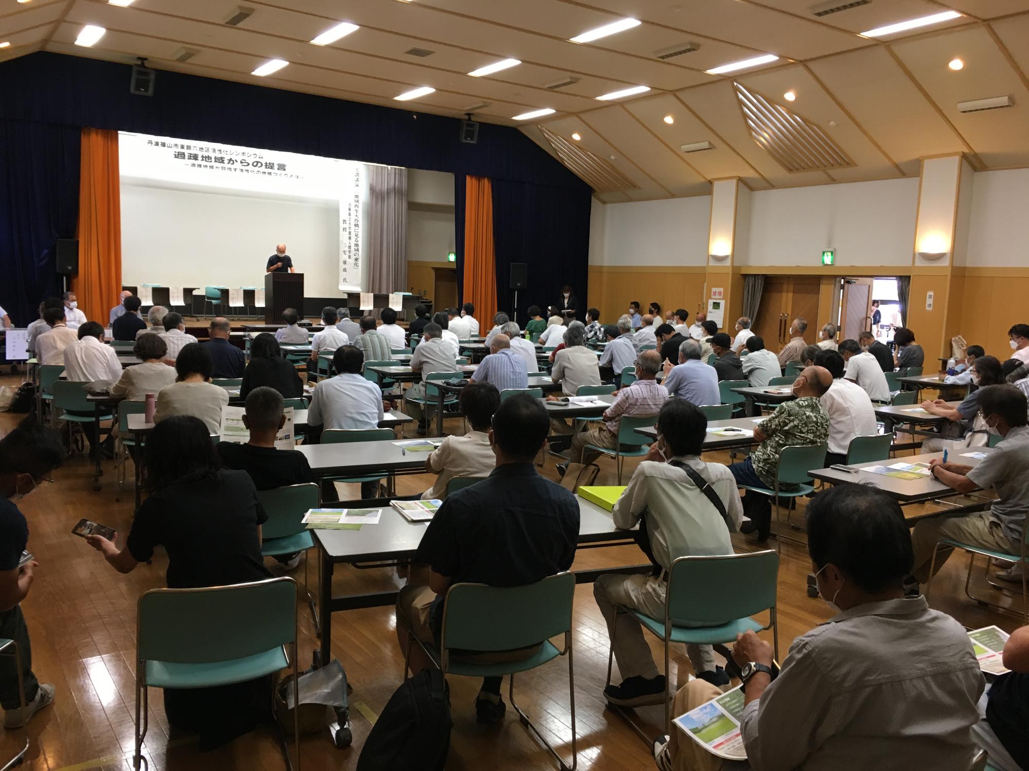 会議室でシンポジウムが行われていて、壇上でお話をしている。会場いっぱいに参加者が座っている。