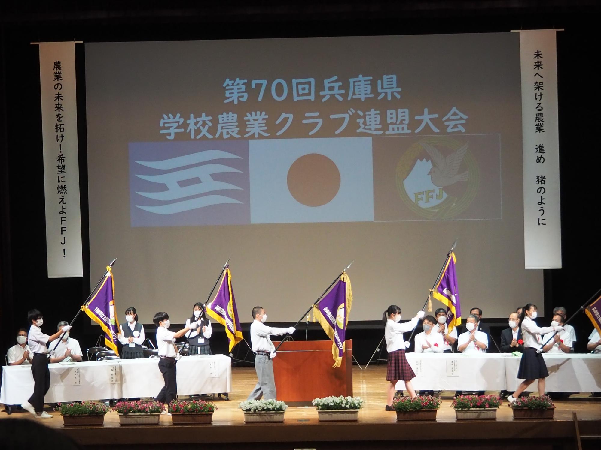 農業クラブ連盟の大会で、舞台上に関係者や来賓の方が並んで座っていて、その前を旗を持った旗手が退場している