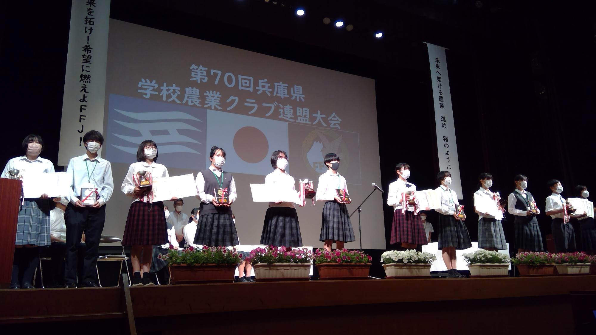 舞台上に、表彰状や盾を持った学生たちが並んで立っている