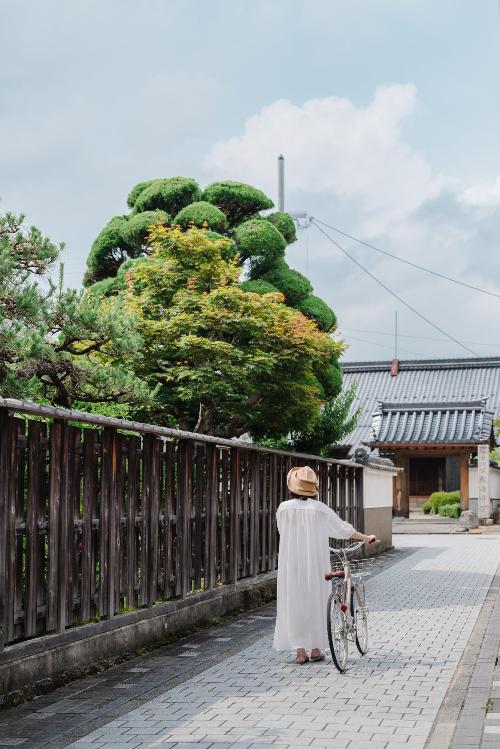 お寺の前を、女性が自転車を押しながら歩いている