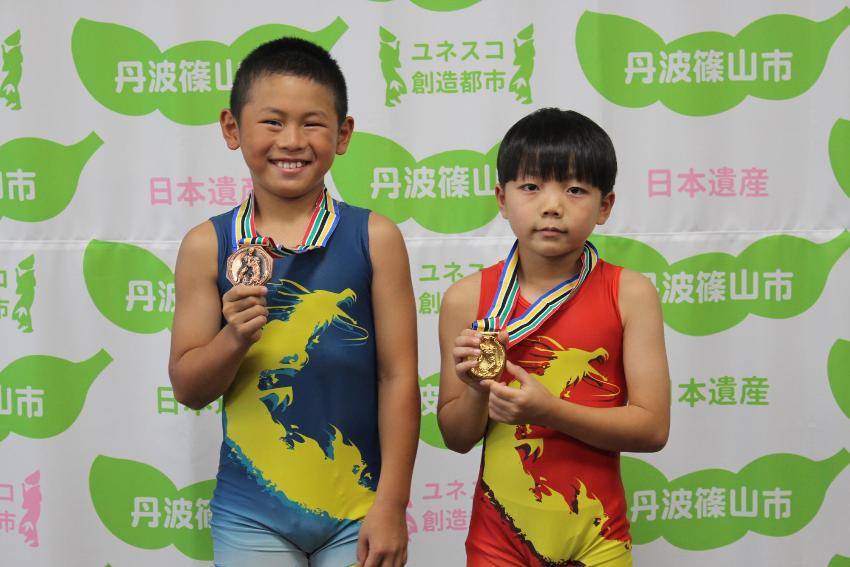 レスリングのユニフォームを着た男の子2人が、首から下げたメダルを手に持って記念撮影