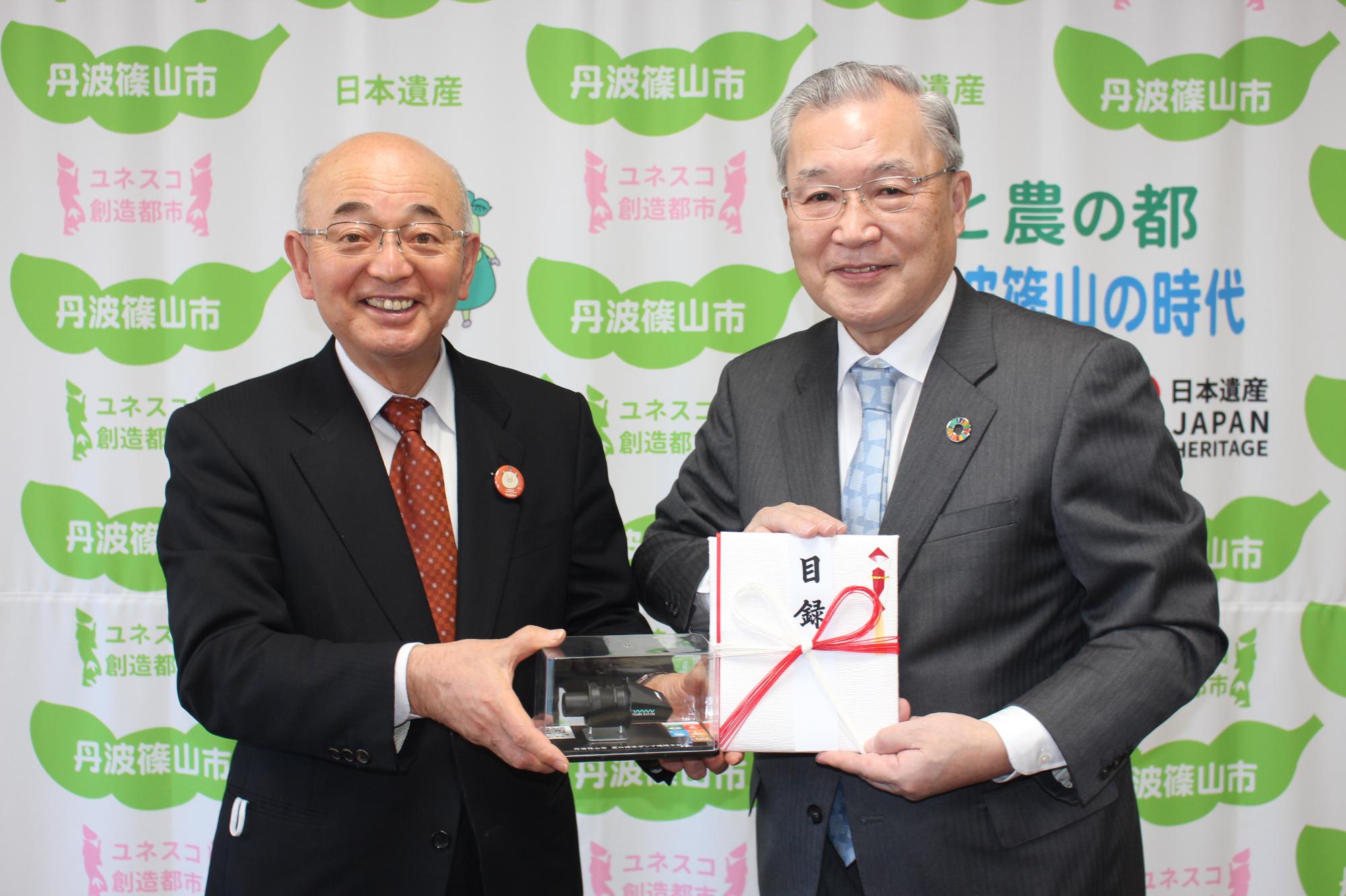 市長が配水ポンプの模型を手に持ち、企業の方が寄付目録を持っている。