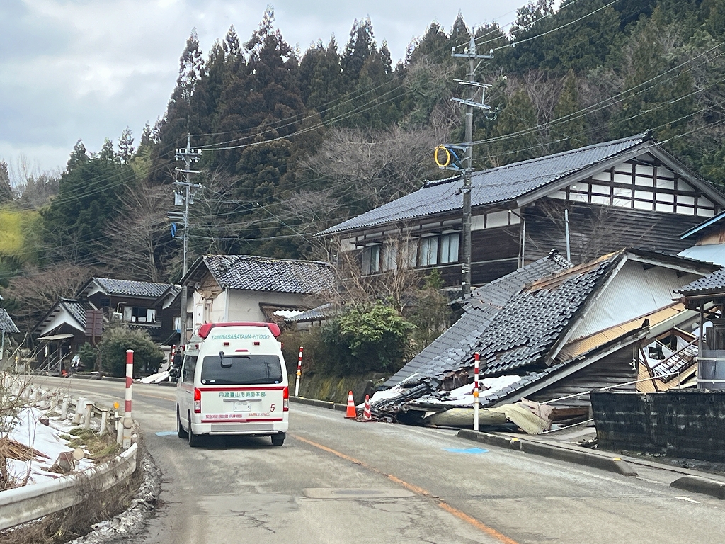 輪島市を道路を走る丹波篠山市の救急車。周りの家が倒壊している。