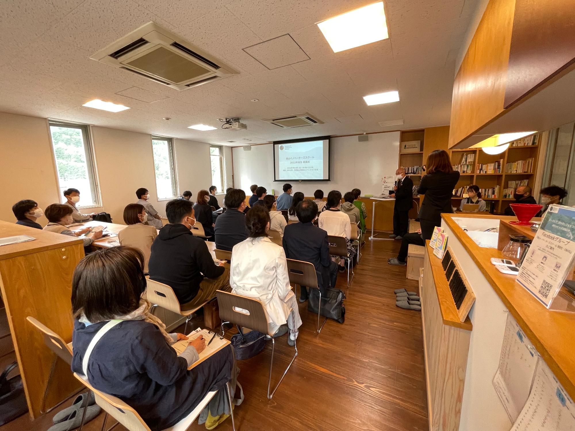 会議室で受講生が椅子に座り、市長が立ってあいさつしている