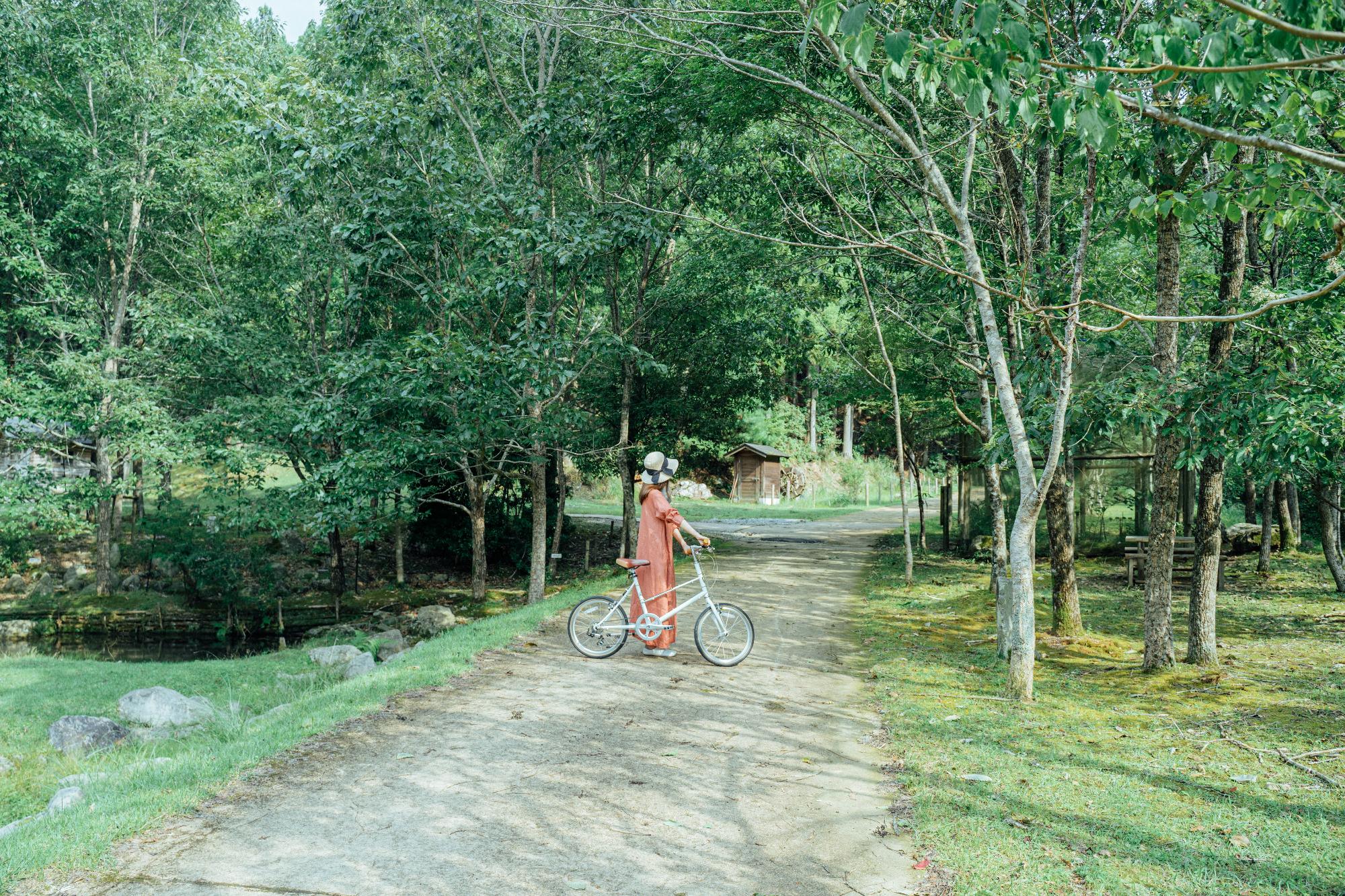 ささやまの森公園内の道で、帽子をかぶった女性が自転車を降りて止まっている