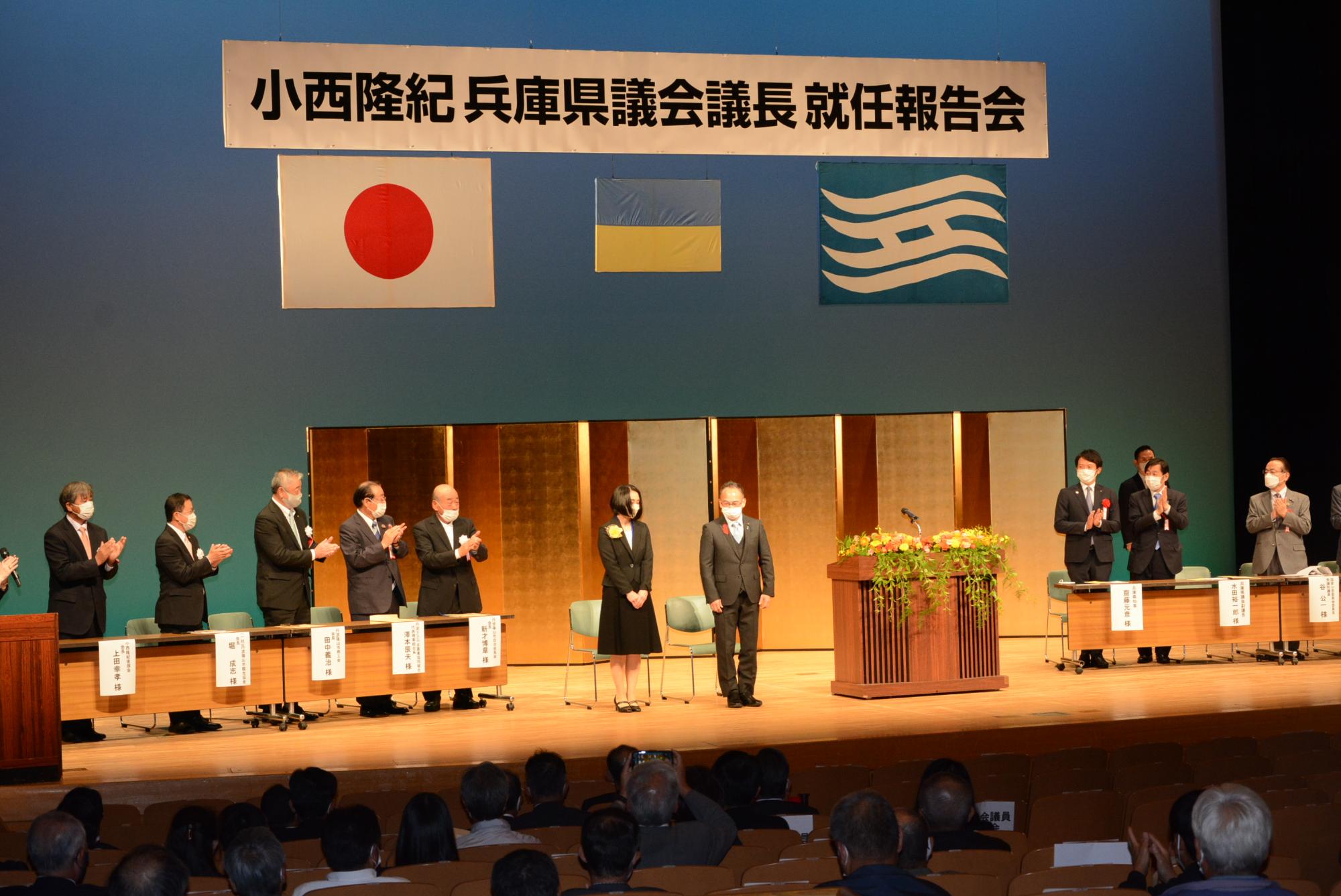 小西議長の就任報告会で議長がステージの真ん中に立って拍手を受けている