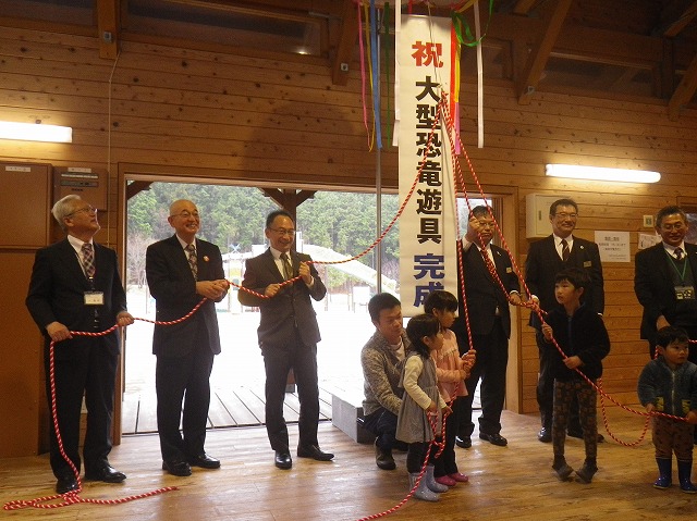 恐竜遊具の完成式典で関係者や子どもたちがくす玉をわっている