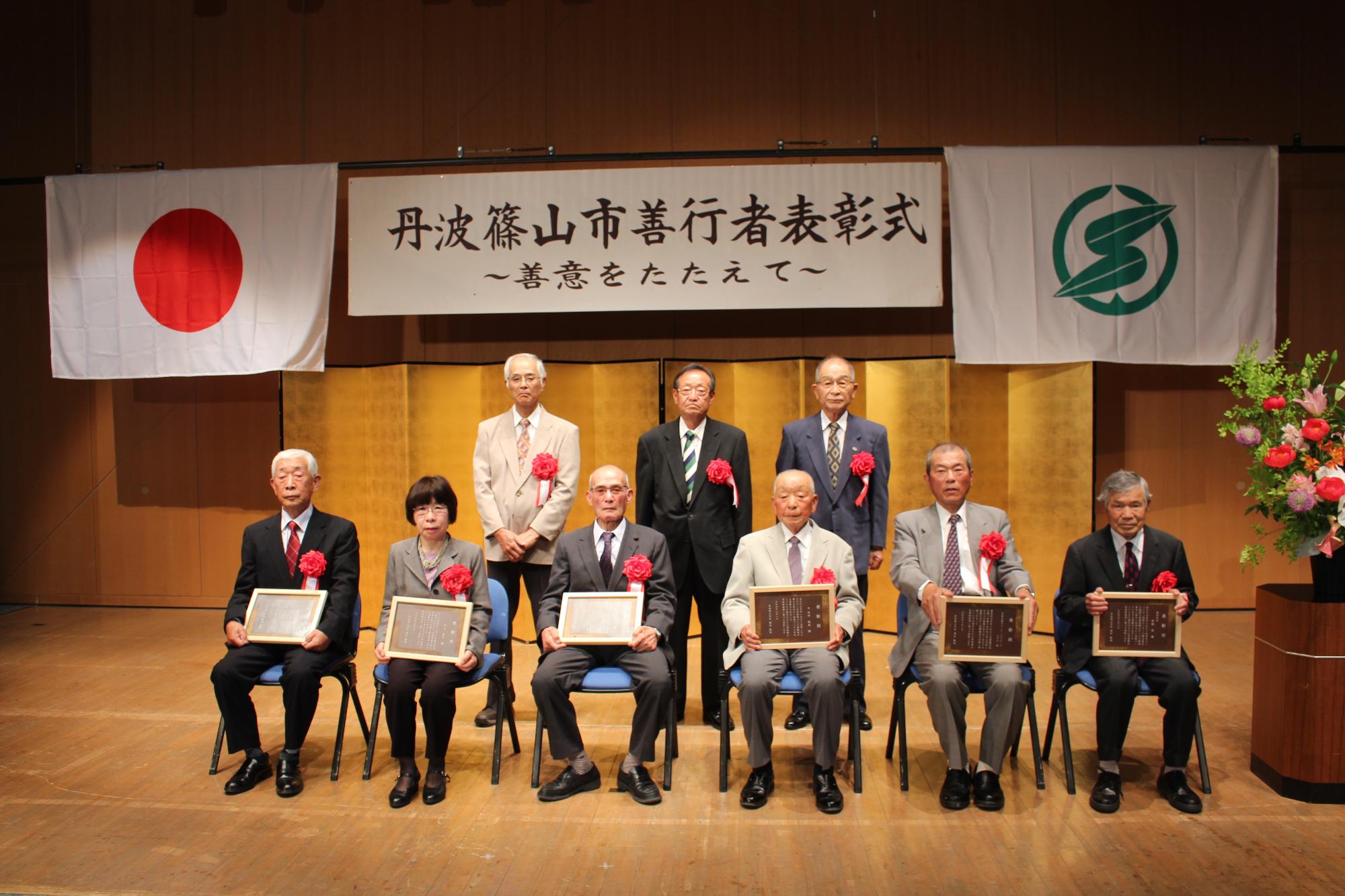 市の善行者表彰式で、前列に表彰盾を持った受賞者の方たちが椅子に座っていて、後列にも受賞者の方3名が立っている。