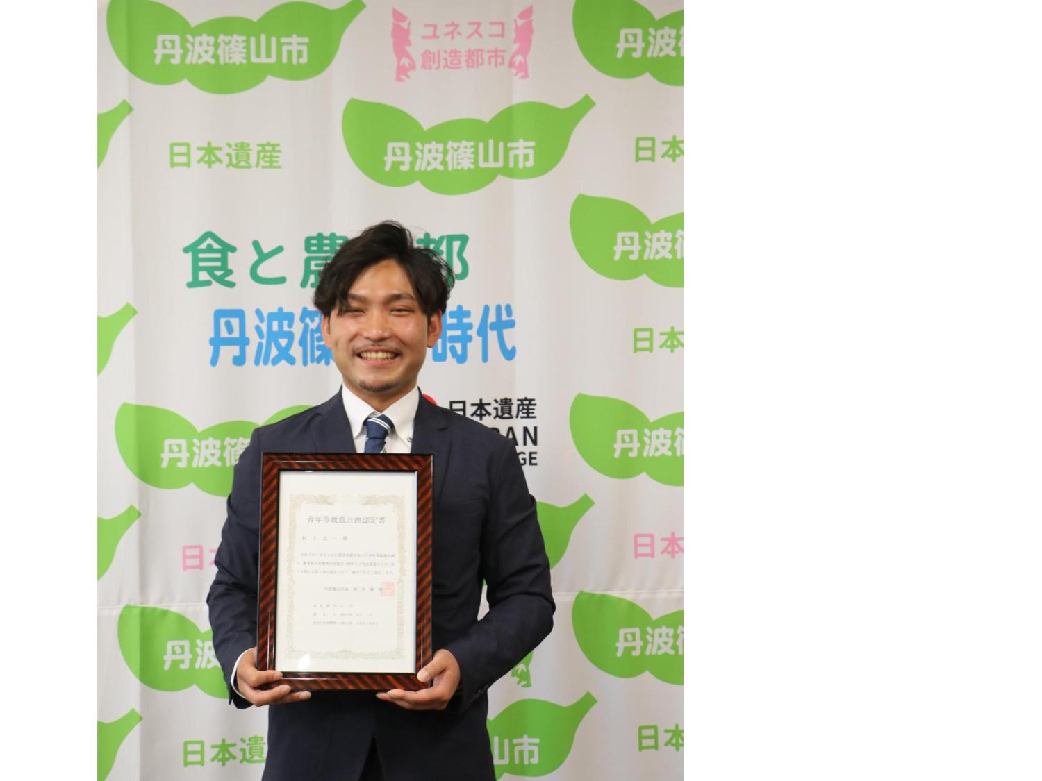 スーツ姿の村上さんが認定証を持っている