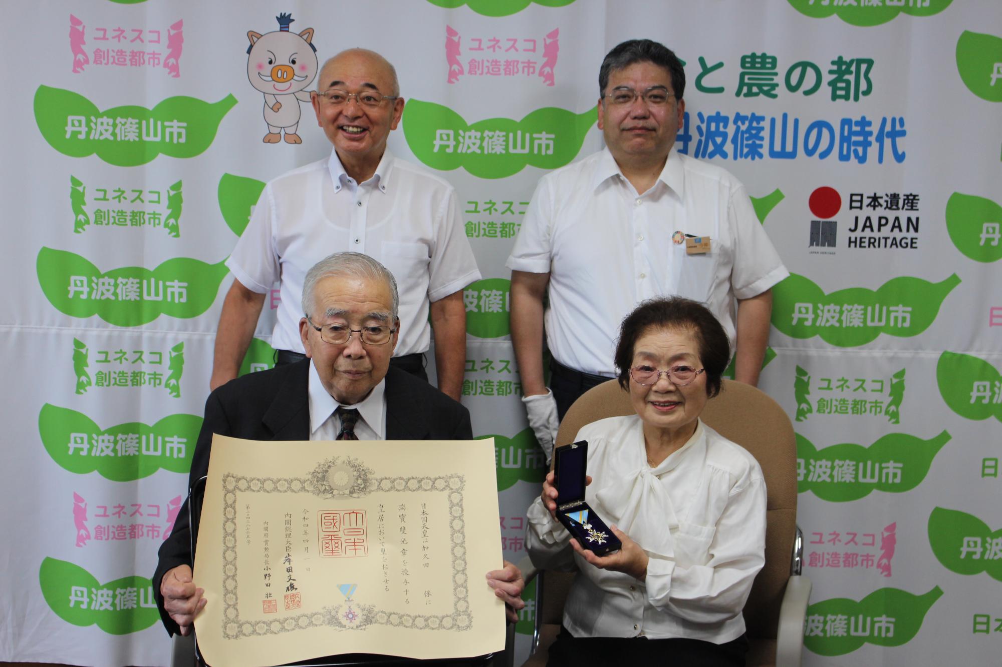 高齢者叙勲の賞状を持った男性、隣に奥様が勲章を持って座っている。後方に市長と県民局長が立っている。