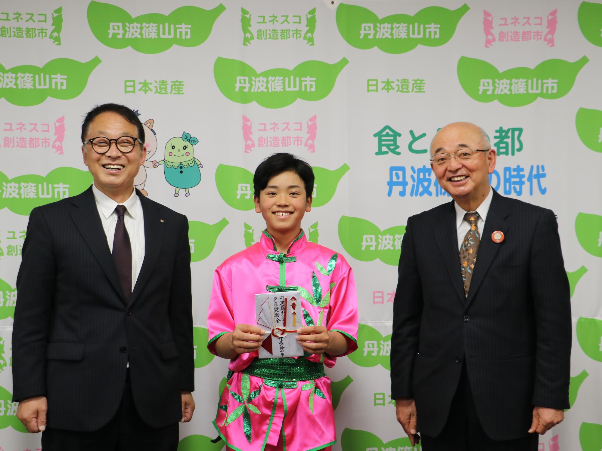 ピンク色の衣装を着た男の子が奨励金の目録を手に持って、両隣に市長と教育長