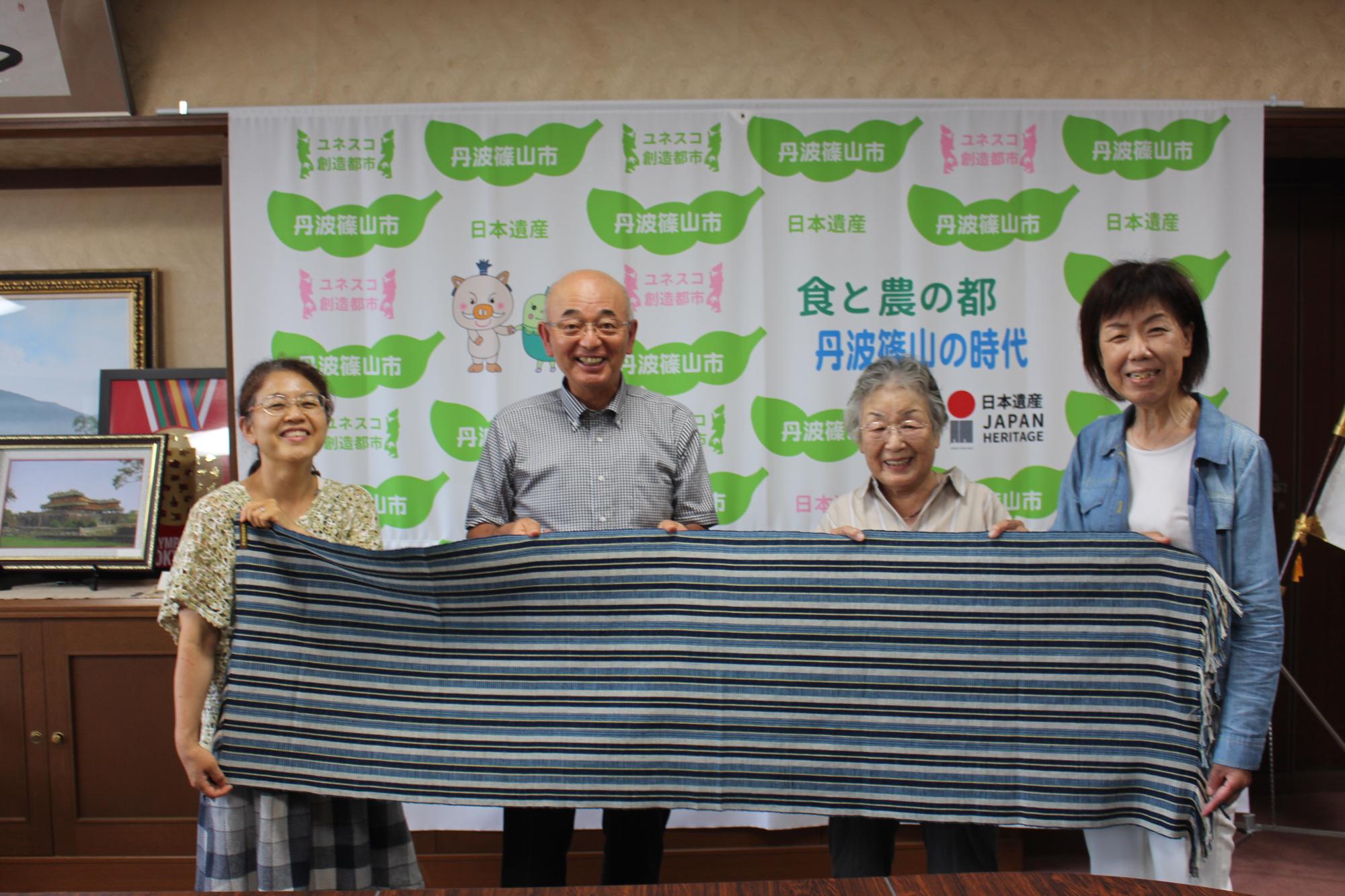 丹波木綿の敷物（テーブルセンター）を、製作された女性3人と市長が広げて持っている